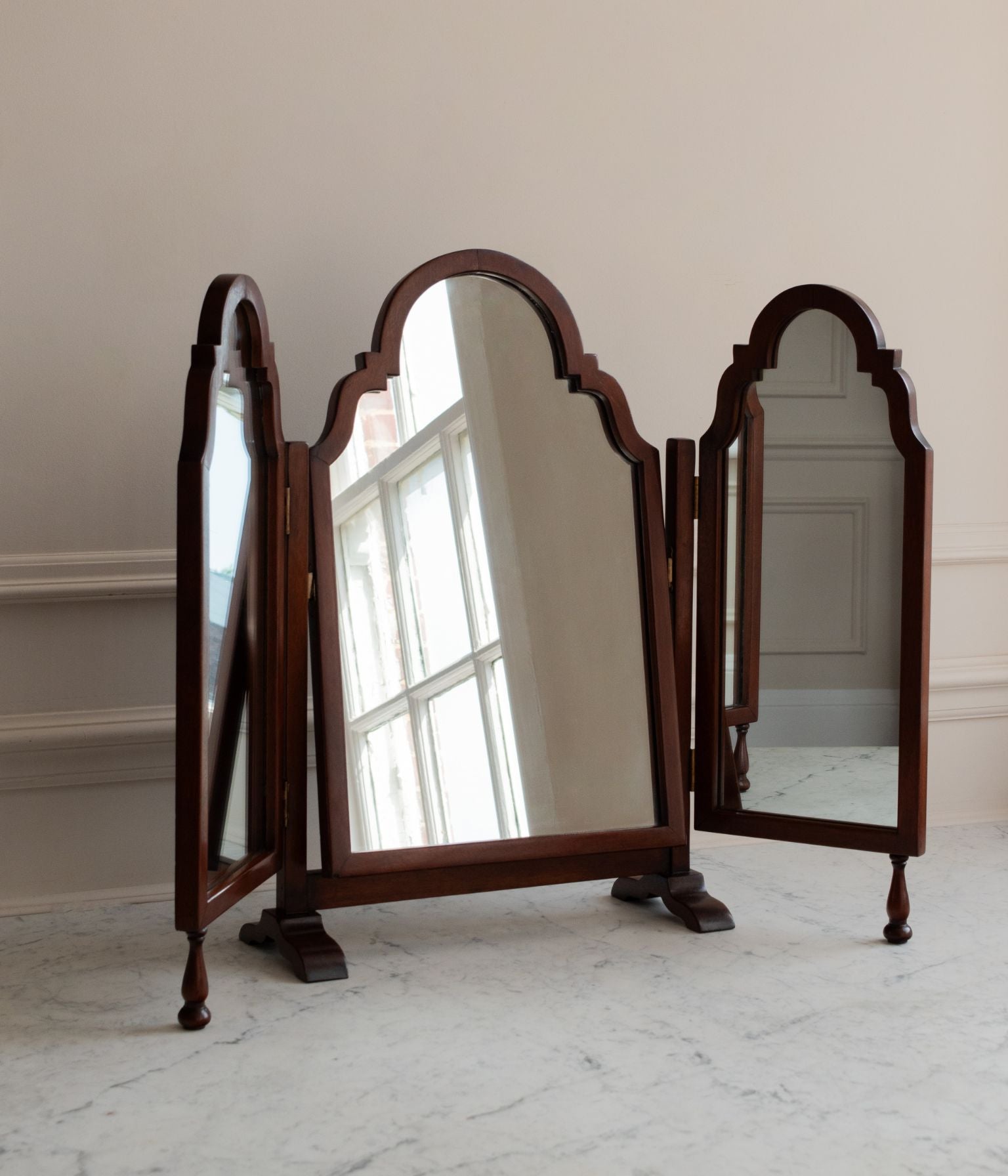 A three-panel wooden dressing mirror with arched tops and turned feet, shown open on a light marble floor against a cream wall.